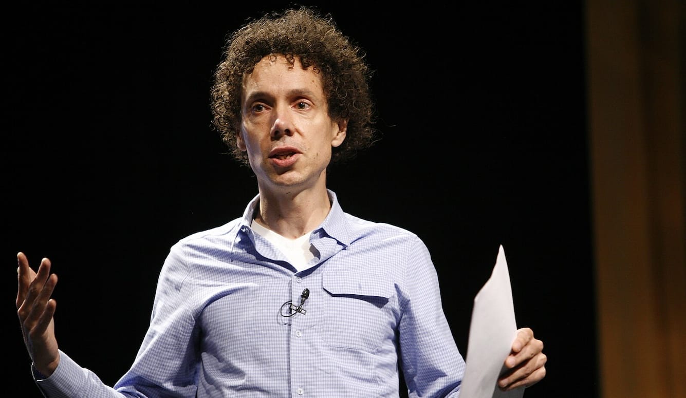 Malcolm Gladwell is a middle-aged white man with curly hair. He is on stage, talking, holding a piece of paper.