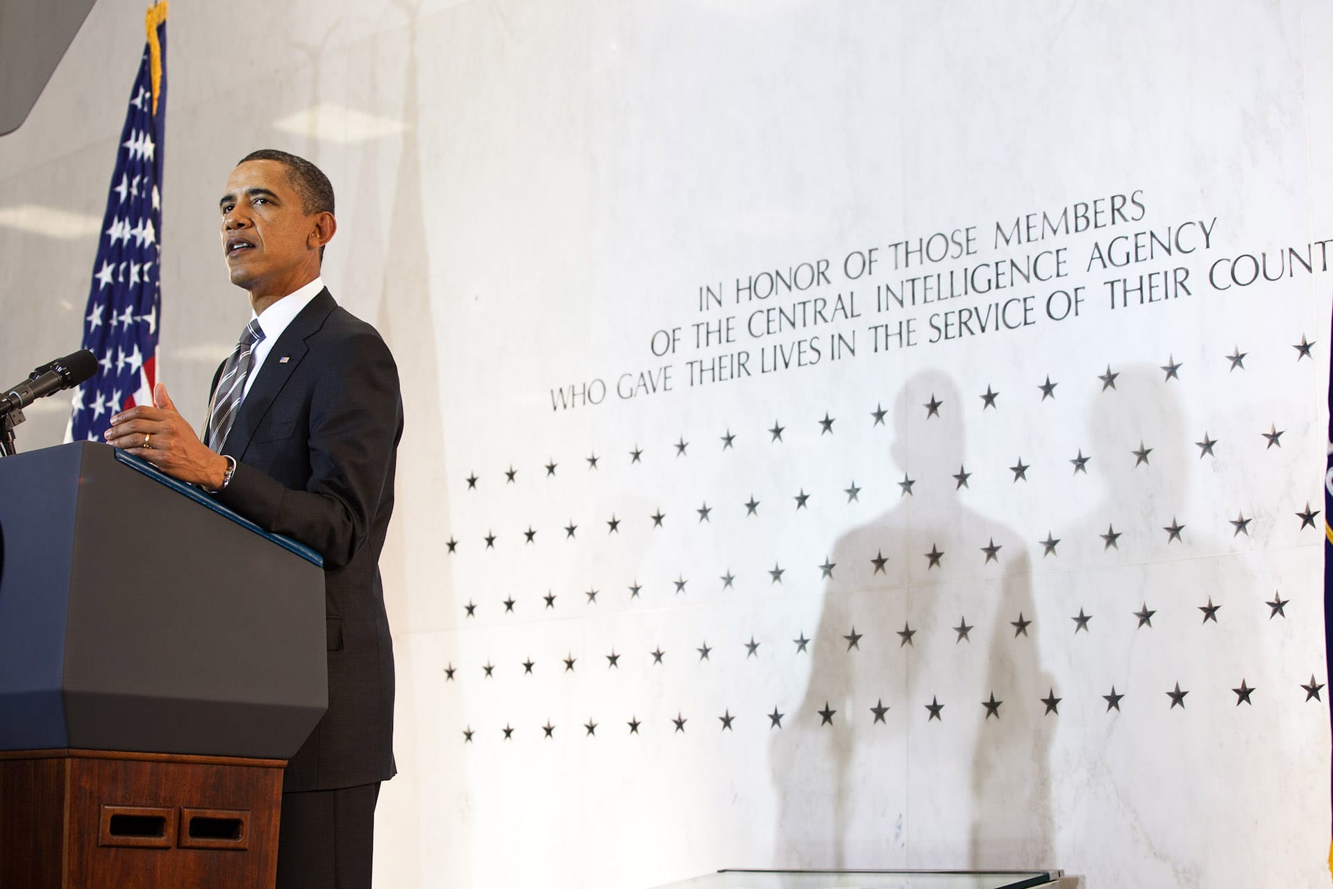 Barack Obama wears a suit and speaks at a podium in front of the CIA wall of stars.