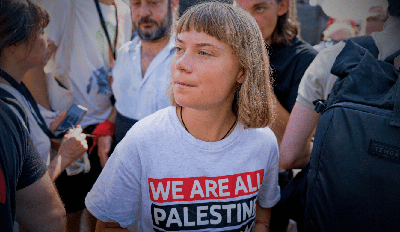 Greta Thunberg is a young white woman with short blonde hair and short fringe. Her shirt says WE ARE ALL PALESTINE.