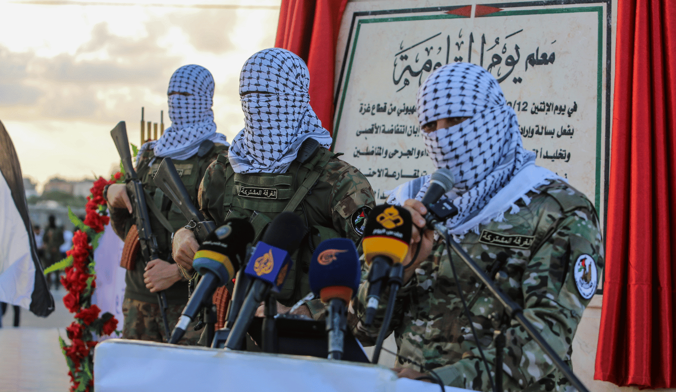 Three masked men in keffiyeh camouflage stand at a podium with rifles and multiple microphones during a press event in Gaza.