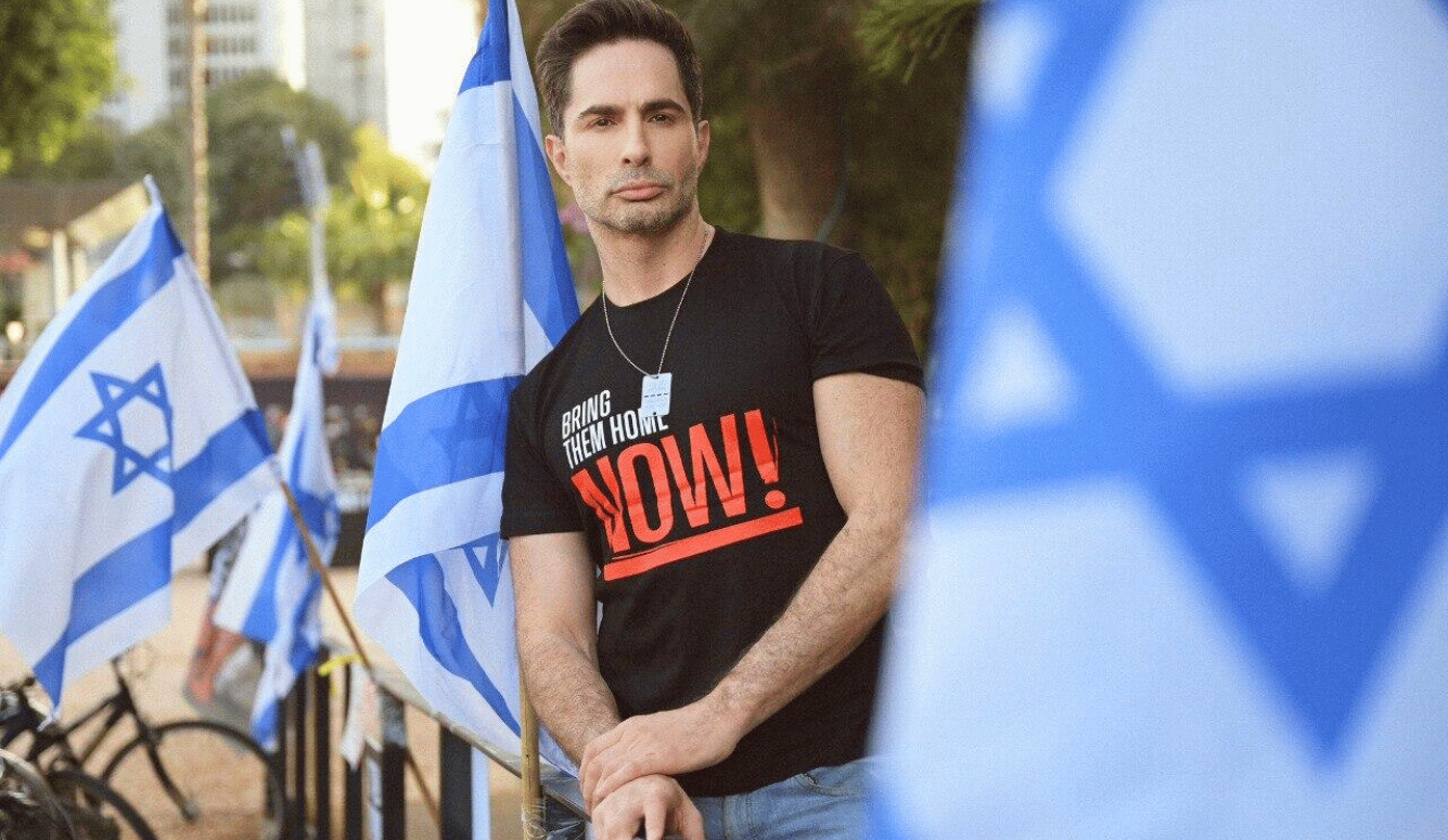 Michael Lucas wearing a “Bring Them Home Now!” T-shirt stands among Israeli flags at a demonstration.