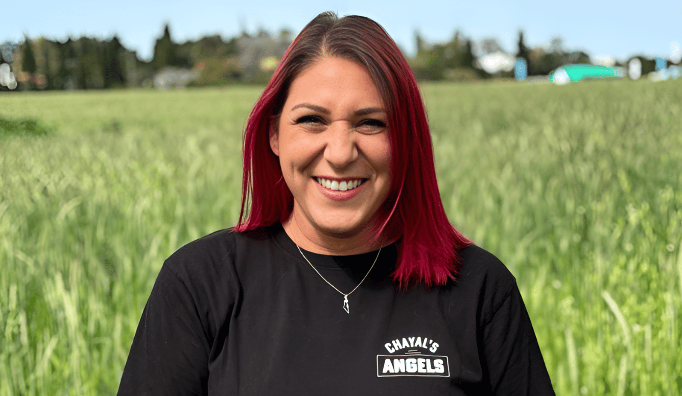 Tasha has bright red (dyed) hair and she is smiling in front of grass paddock. She wears Chayal's Angels tshirt.