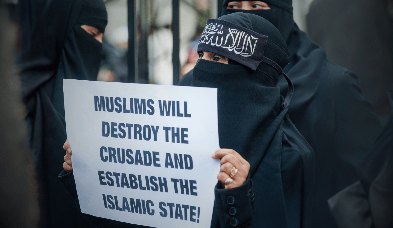 Woman in black niqab holding a protest sign reading “Muslims will destroy the crusade and establish the Islamic State.”