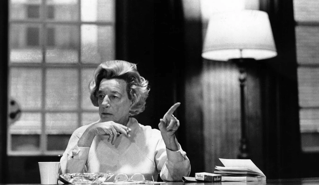 Black-and-white photo of a Lillian (an elferly woman) seated at a table, gesturing with one hand beside papers, glasses, and a coffee cup.