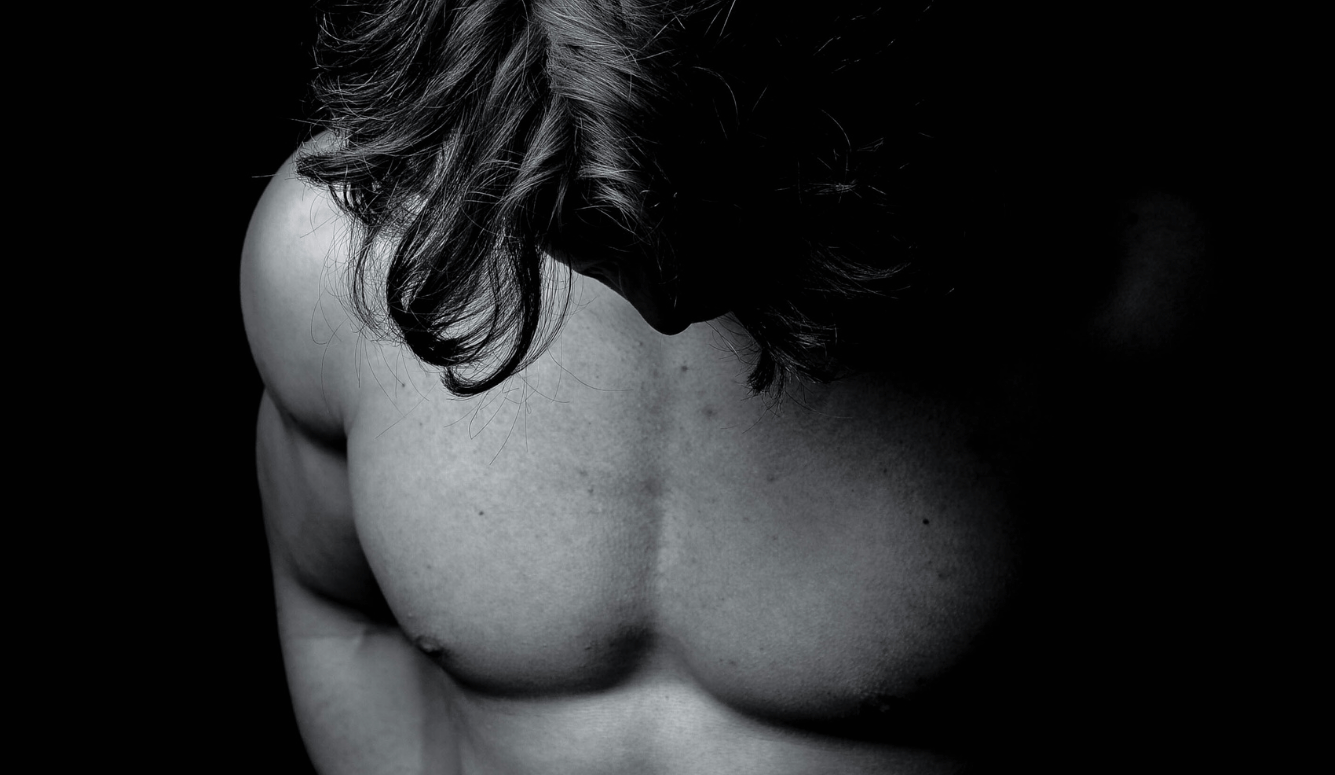 Topless young man with long hair looking down, showing defined upper torso in low light.