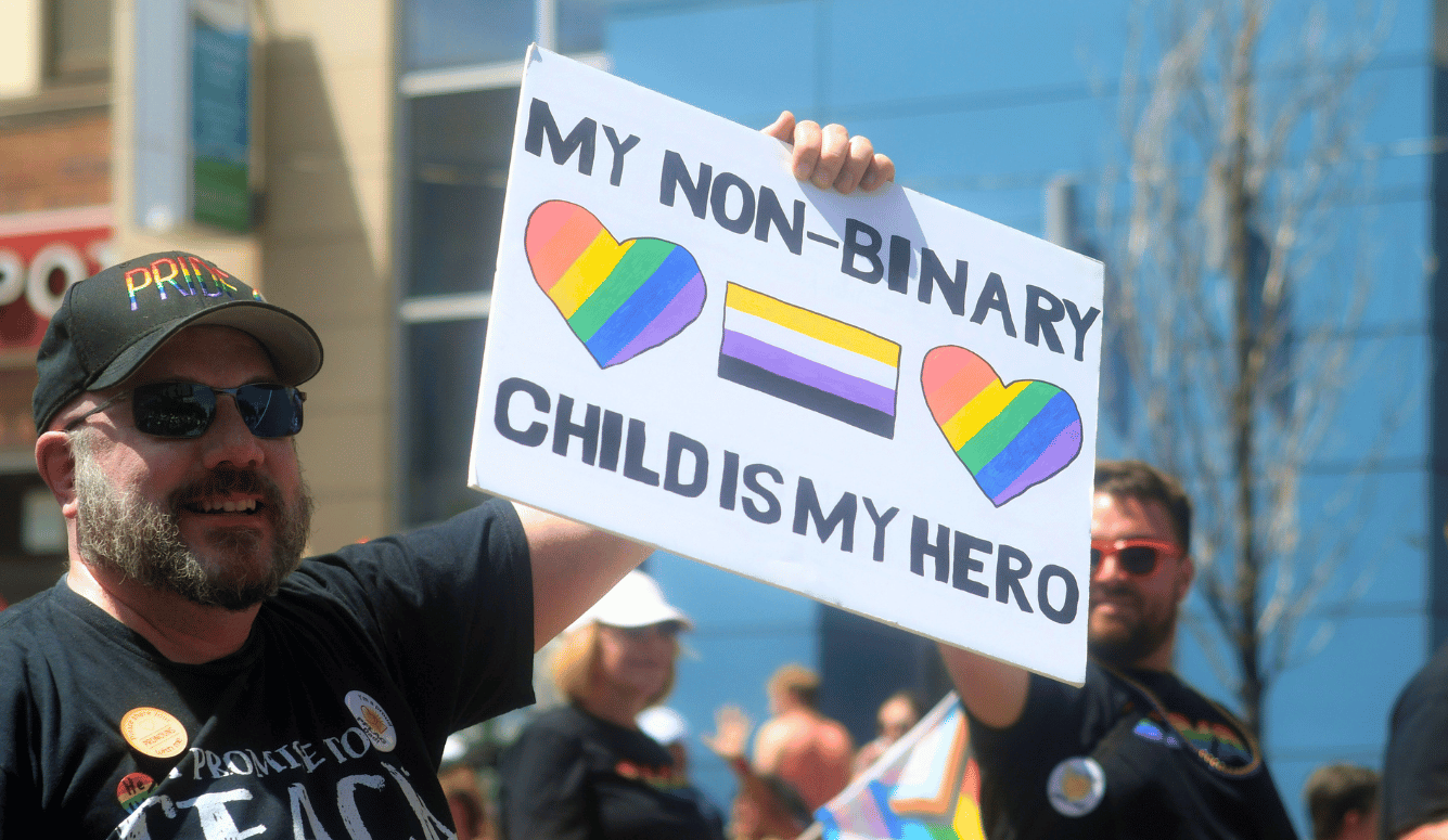 Man in sunglasses and a Pride cap holds a sign reading “My non-binary child is my hero” at an outdoor parade.