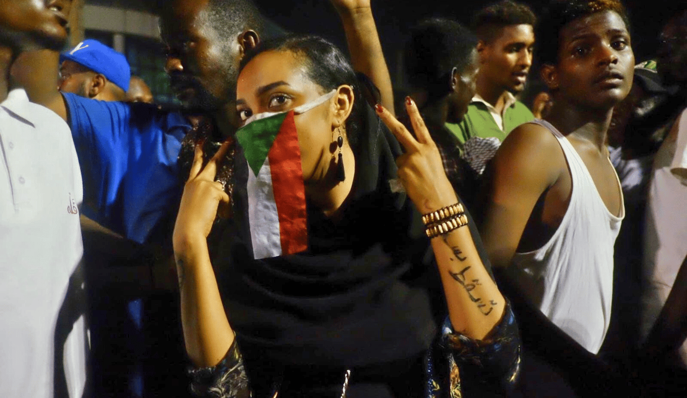A woman wearing a Sudanese flag face covering makes a peace gesture during the 2019 Khartoum sit-in, with script on her arm calling for the government to fall.