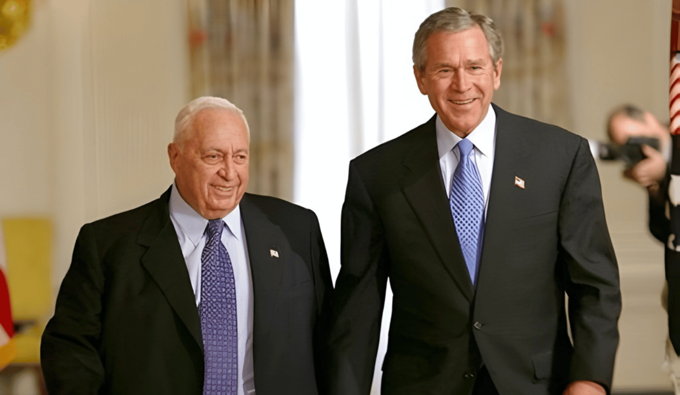 US President George W. Bush and Israeli Prime Minister Ariel Sharon prior to talking with the press in the Cross Hall of the White House. They wear suits and look happy.