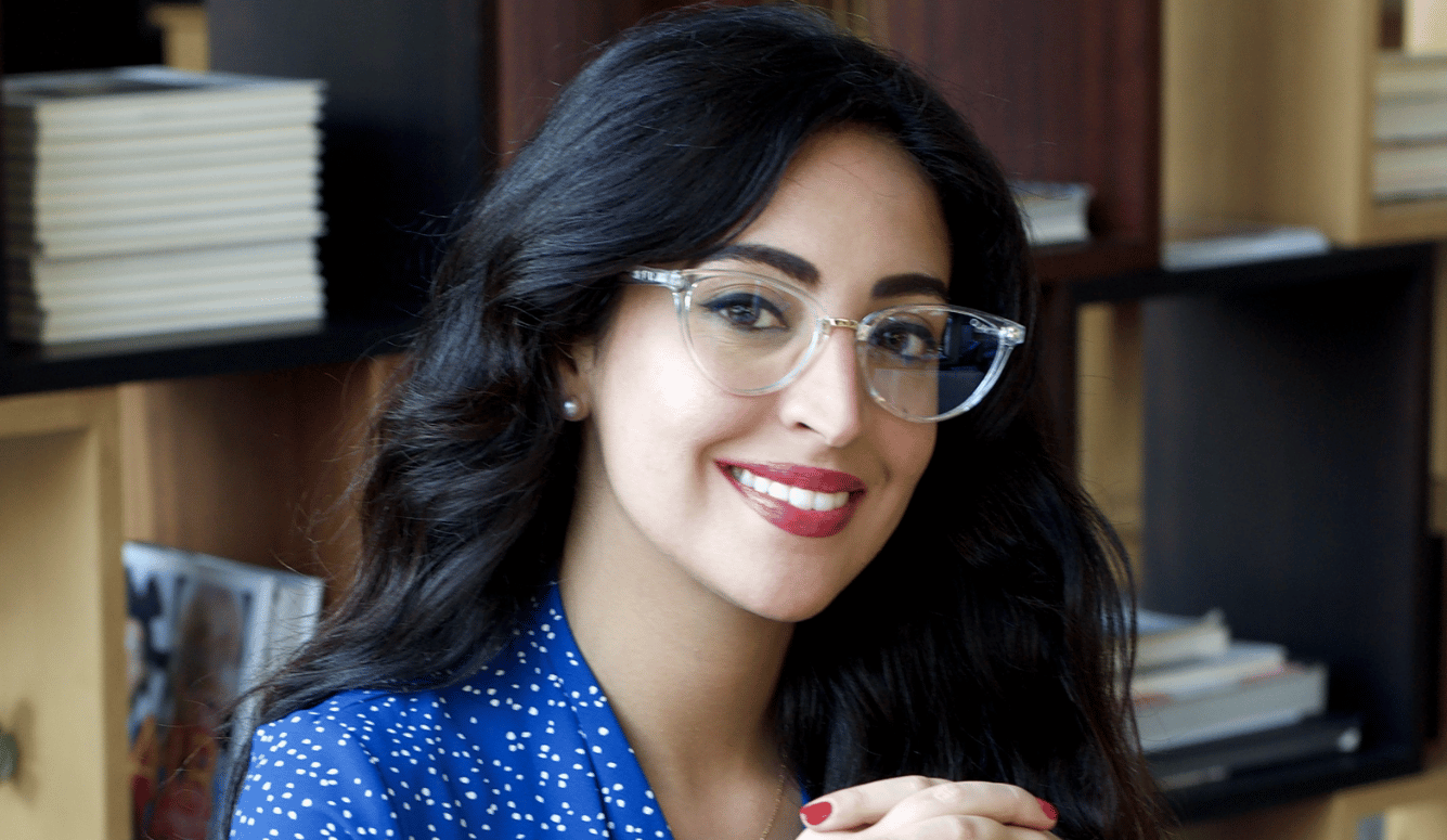 Chama Mechtaly is a young woman with olive skin, long dark hair, and glasses. She sits in front of a bookshelf.