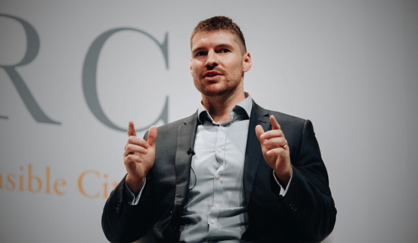 Gerard Holland is a man in his thirties in a suit. He stands on a podium. 