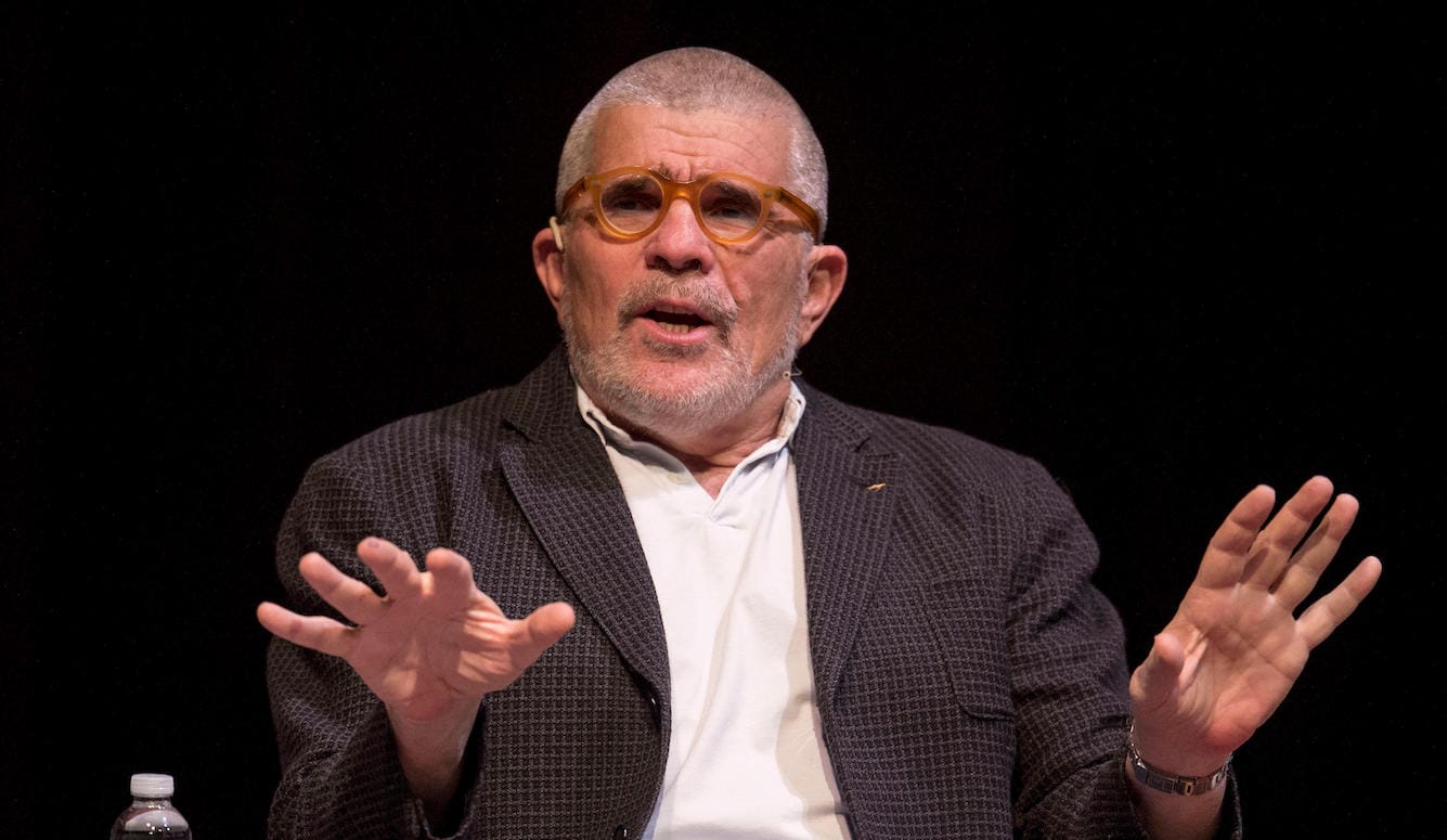 David Mamet is a white man in late middle age. He is talking and gesticulating. He wears glasses, a T-shirt, and a jacket.