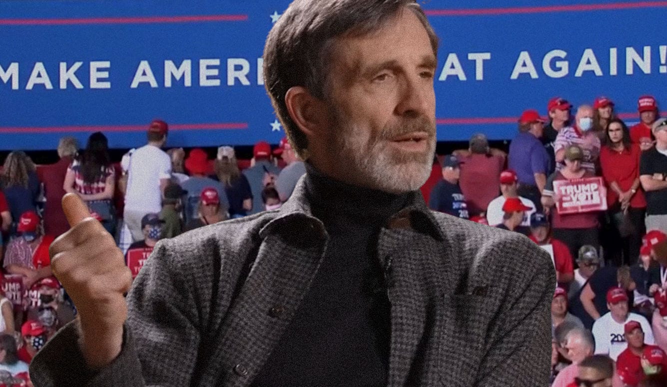 A white man in a check jacket speaking in front of a backdrop of a MAGA rally