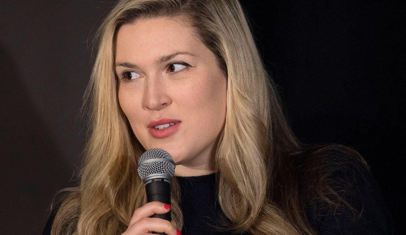 Young blonde woman speaking into a microphone against a dark background.