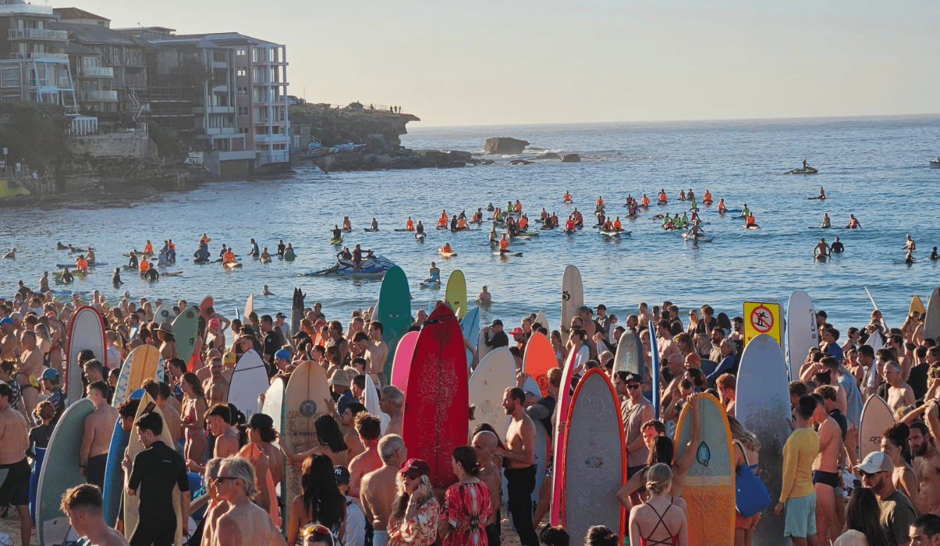 Crowded Bondi Beach with surfers holding colourful surfboards on the sand and in the water in the early morning.