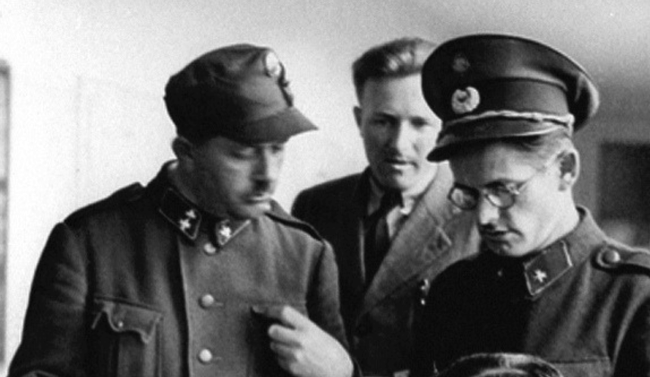 Black-and-white photo of three uniformed SS officers examining documents indoors, wearing peaked caps and military tunics.