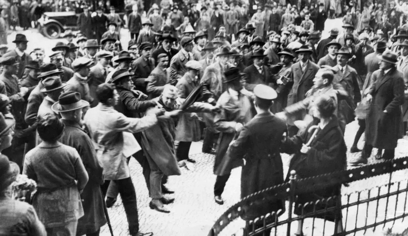 Black-and-white photograph of a crowded street confrontation, with police officers using batons against men in coats and hats.