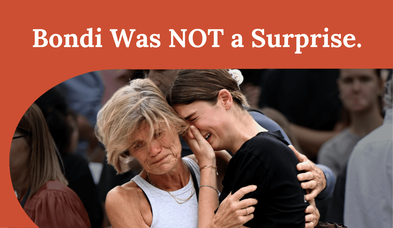 Two women embrace in a crowd, one wiping tears, beneath a banner reading “Bondi Was NOT a Surprise”.