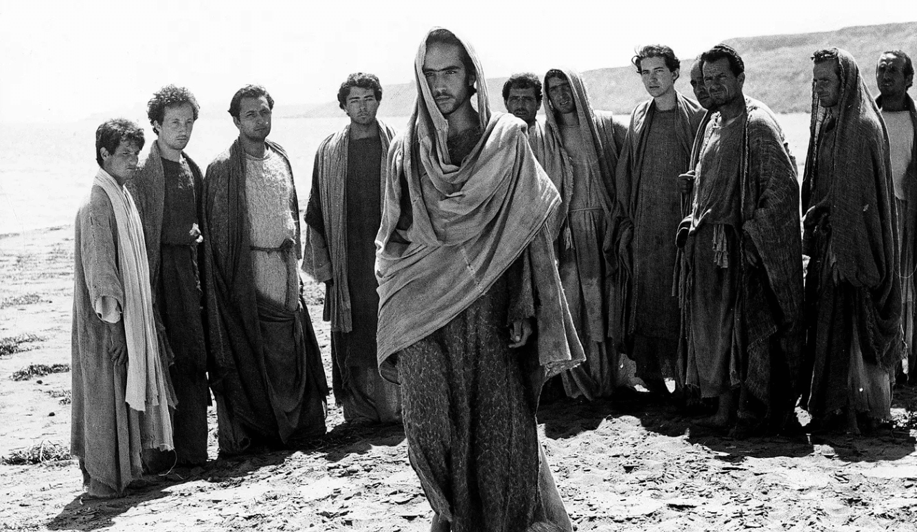 Black-and-white film still showing a robed man walking ahead of a group of men in loose garments in the desert