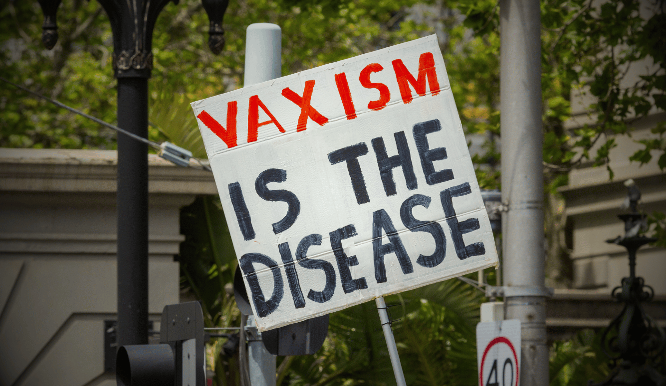 Protest sign reading “Vaxism Is the Disease” held outdoors near street signs and greenery.