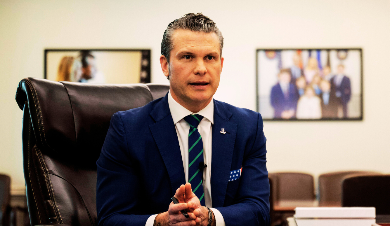 Pete Hegseth at his desk, in a suit and tie. He is a man in his fifties with slicked-back hair. 