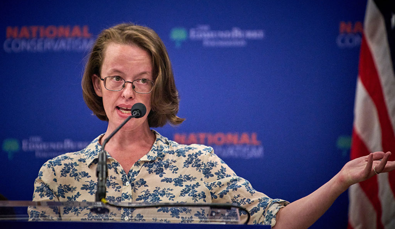 Helen Andrews is a middle-aged woman with short hair and glasses, wearing a floral shirt. She is at a podium.