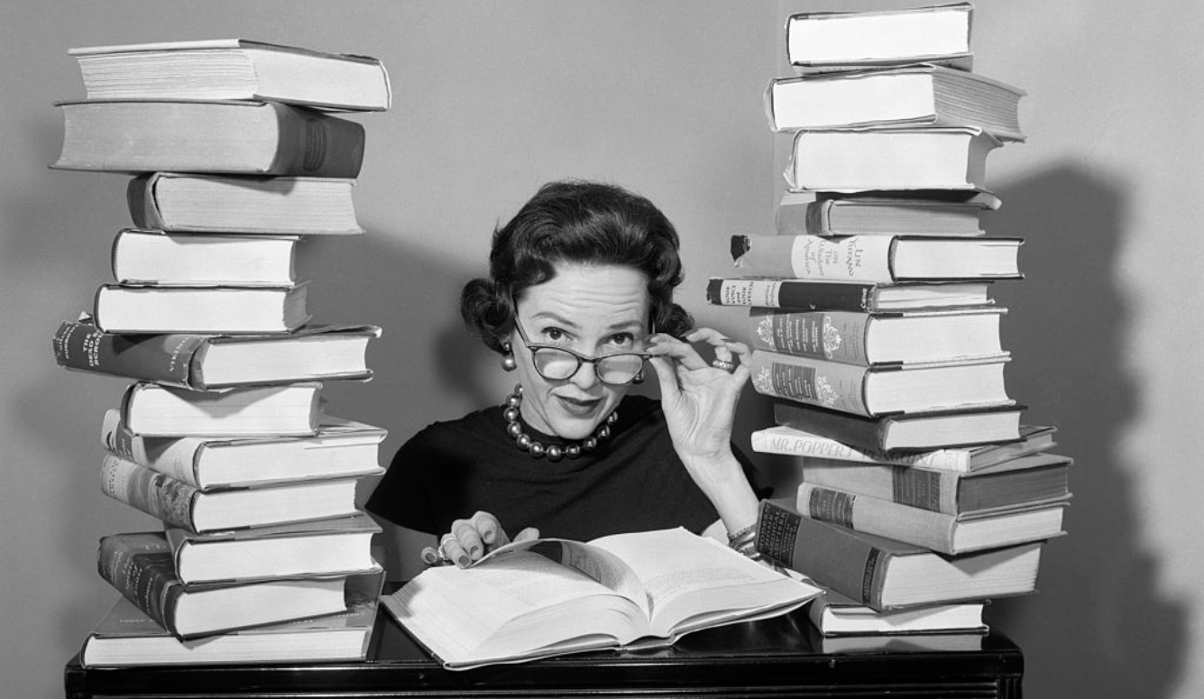 A female librarian with spectacles sitting among a pile of books. 