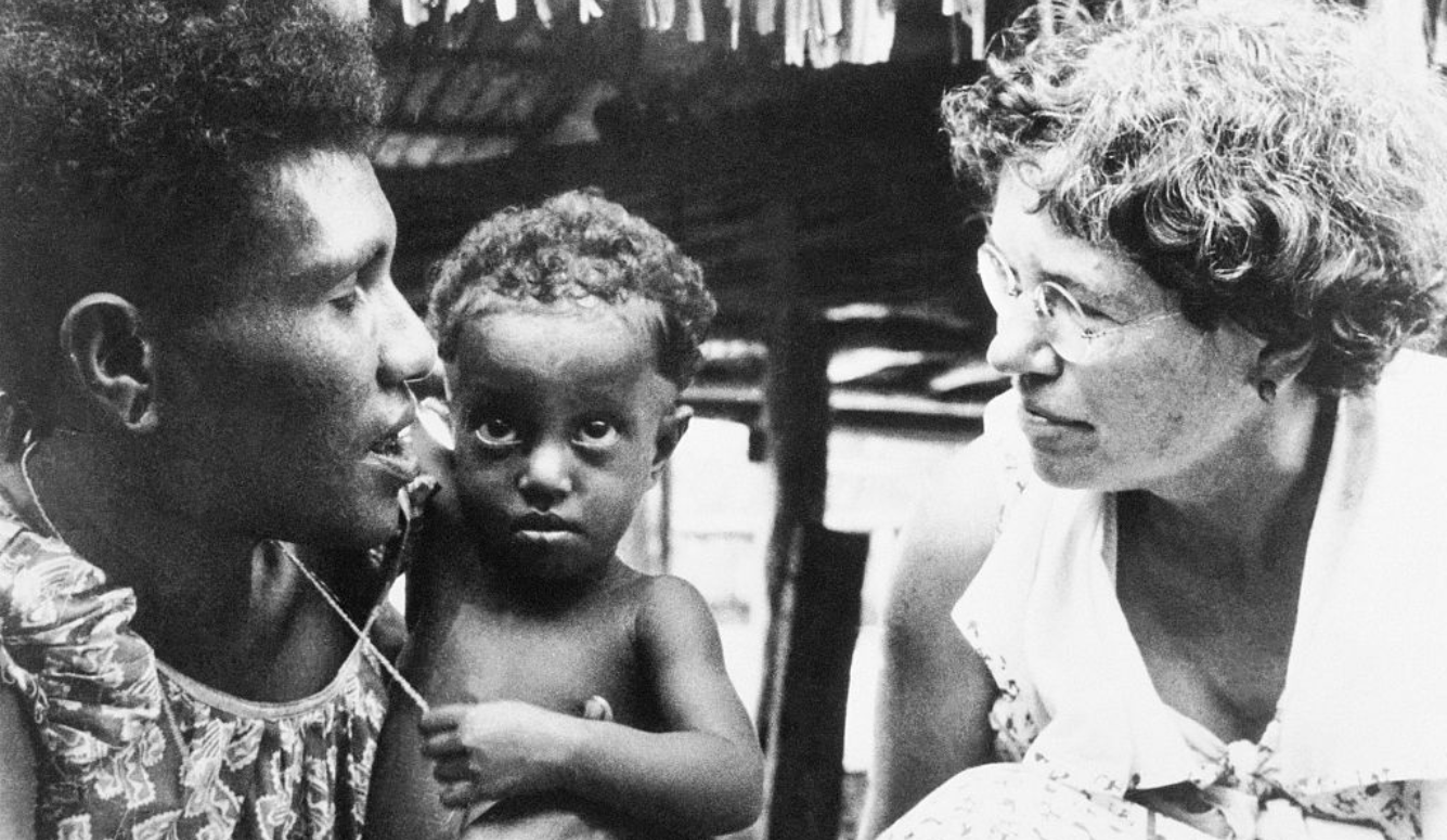 Margaret Mead, a white woman with short hair and glasses, speaks to a dark-skinned mother and child. 