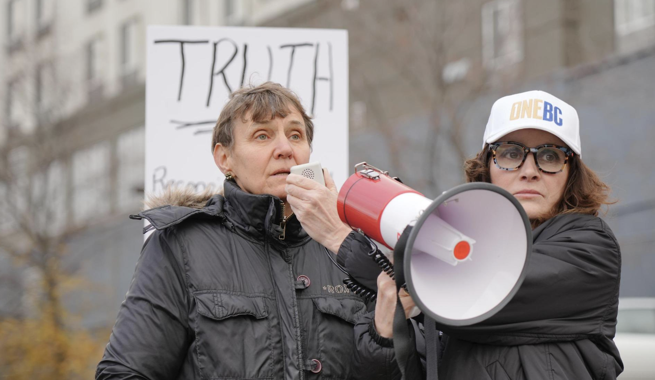 Two women in black rainjackets carrying a megaphone in front of a placard that says Truth. 