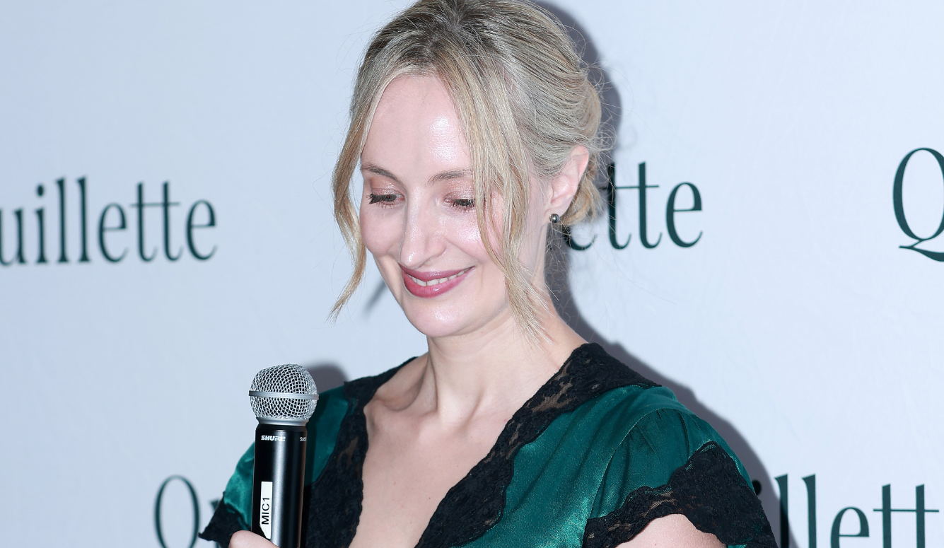 Claire Lehmann is a 40-year-old woman with blonde hair and pale skin. She stands at a microphone, smiling. 