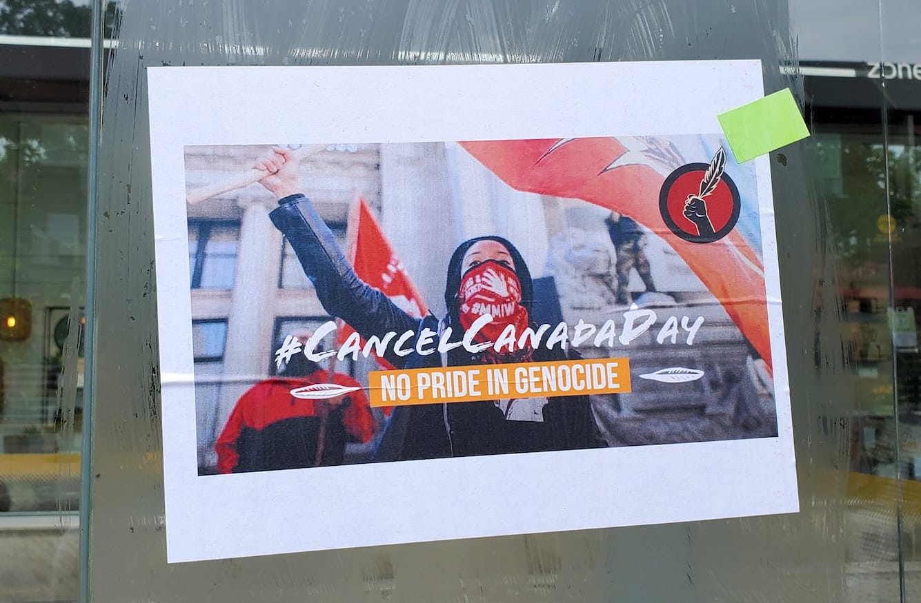 A poster affixed to glass shows a masked protestor. It reads #Cancel Canada Day and No Pride in Genocide. 