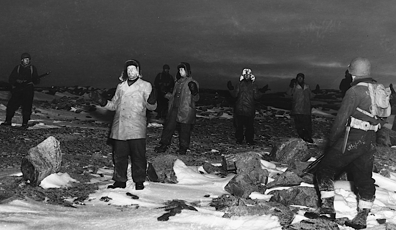 Black and white photograph showing people in heavy winter clothing standing in a snow-patched landscape with debris scattered on the ground.