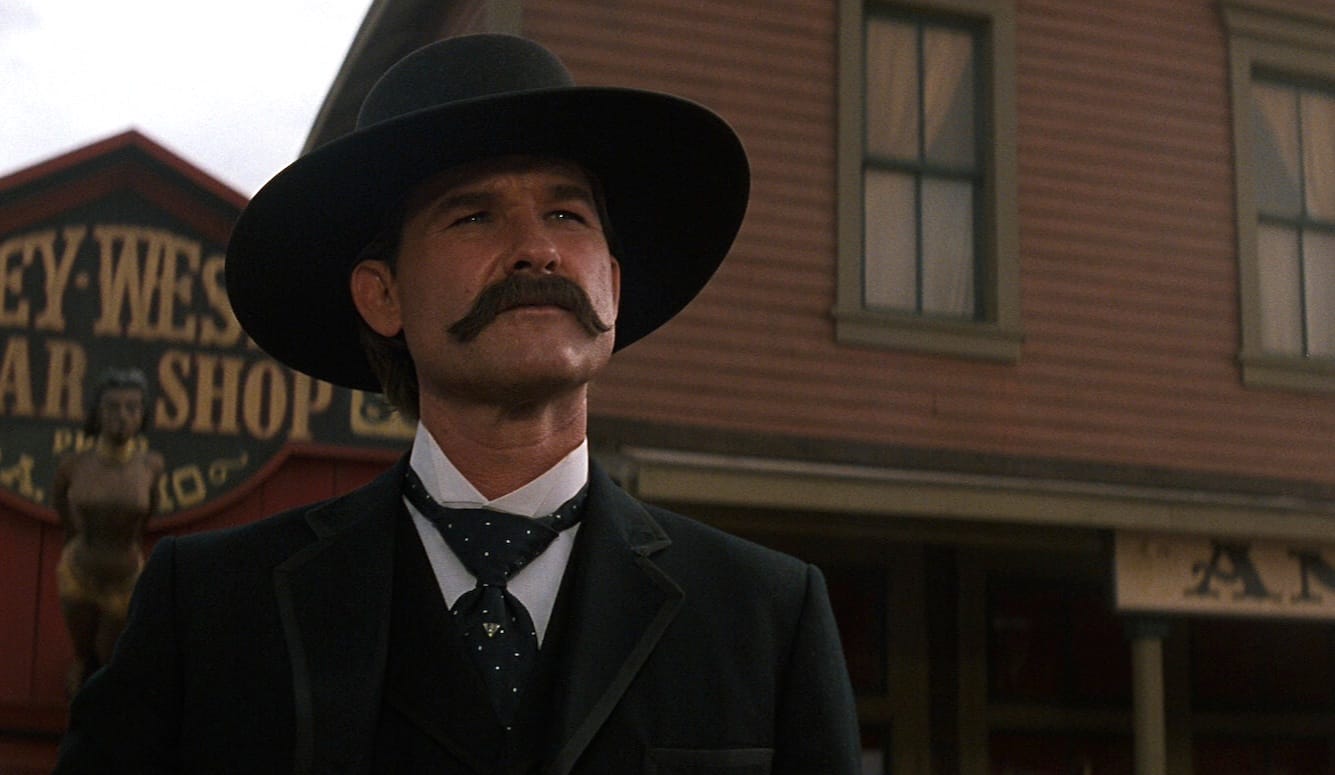Kurt Russell as Wyatt Earp, with a handlebar moustache, jacket, cravat, and hat, in front of a set of a Western saloon. 