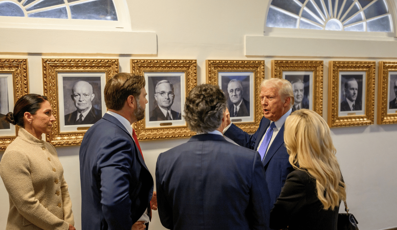 Group of formally dressed politicians in a hallway with framed presidential portraits, Trump in a blue suit speaking to others.