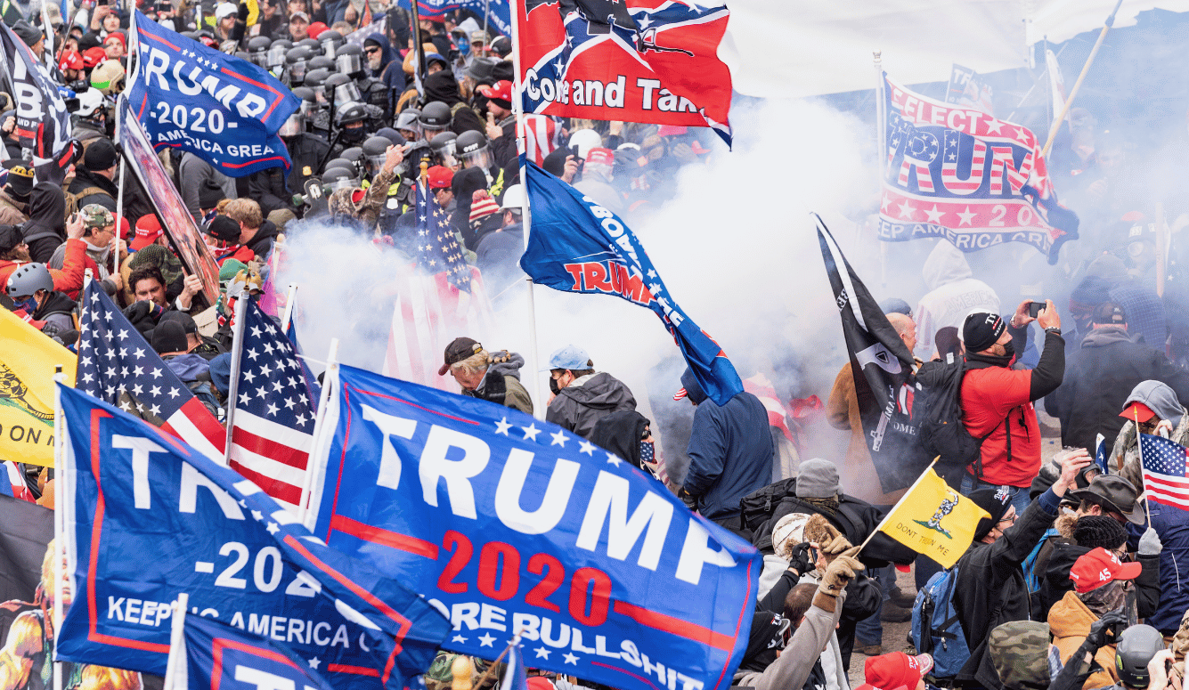 Crowd of protesters waving Trump 2020 flags amid police lines and smoke during unrest outside the US Capitol.