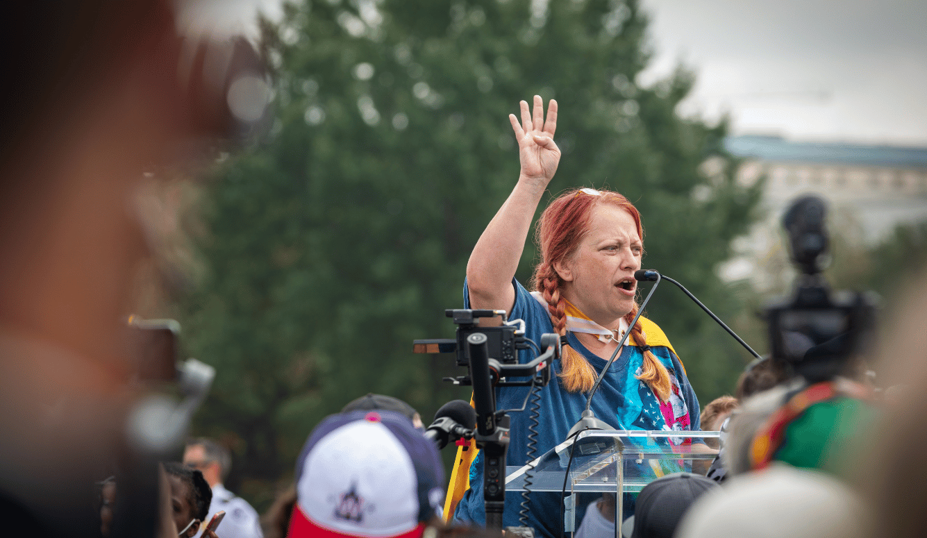 Female speaker with braided hair addresses a crowd from a podium at a Washington DC rally, raising one hand and speaking into a microphone.