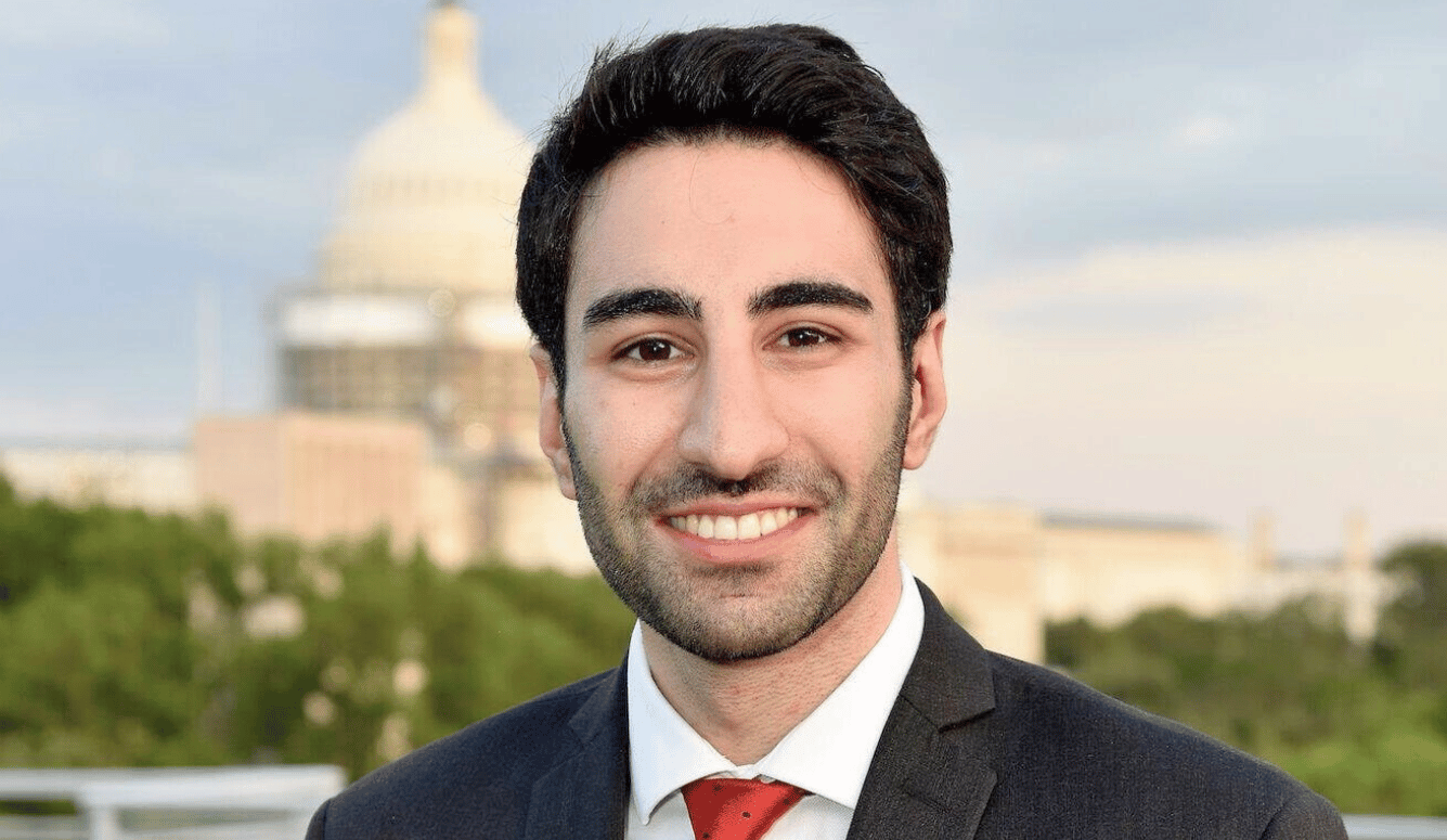 Portrait of a young adult man in a dark suit and red tie, smiling outdoors with the US Capitol dome blurred in the background