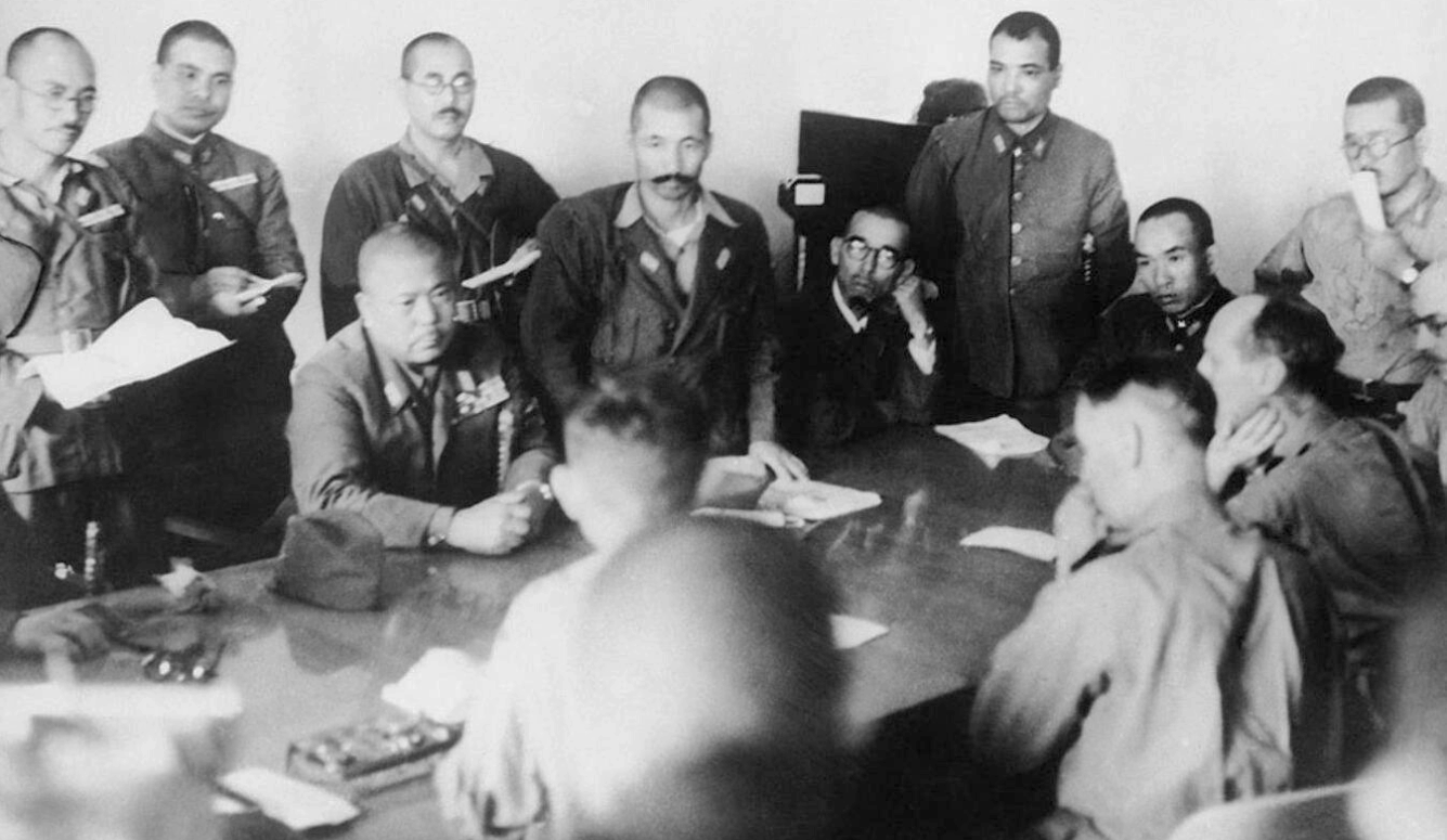 Black-and-white photograph of Japanese military officers gathered around a table during a wartime meeting, with documents and notebooks visible.