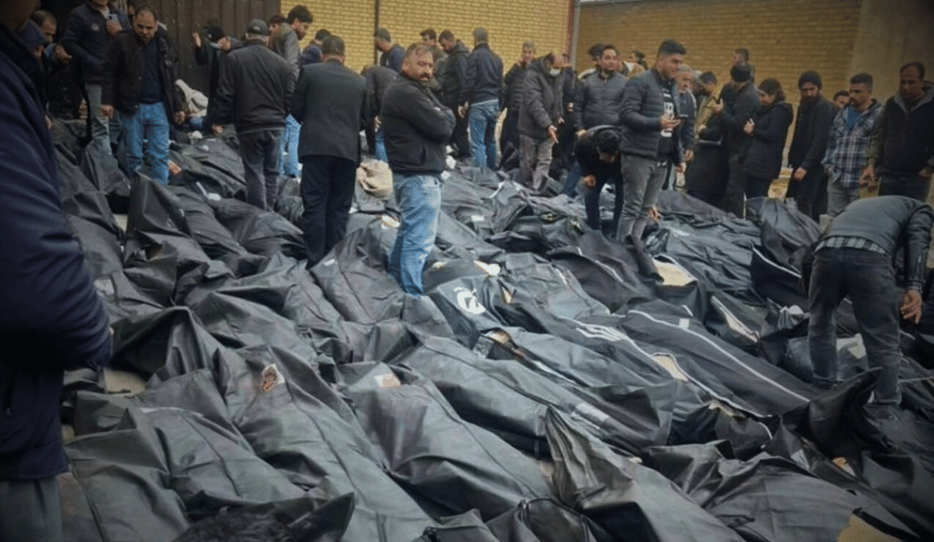 Men stand among rows of black body bags laid on the ground outside a brick building.