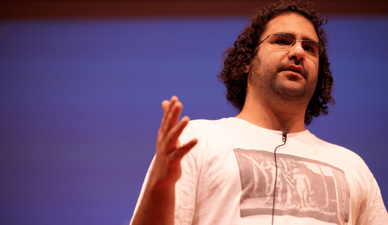 El-Fattah is a man with curly hair and glasses speaks on stage, wearing a white T-shirt and lapel microphone against a blue backdrop.