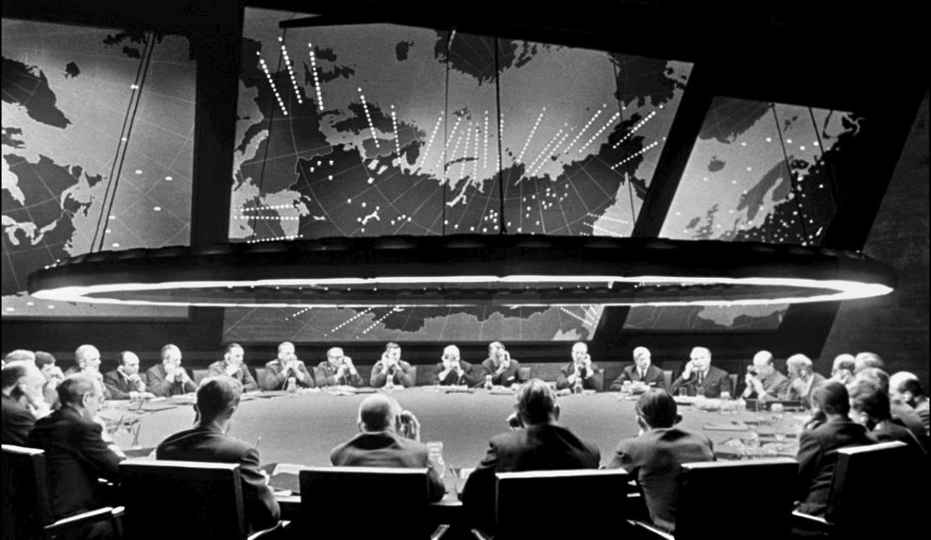 Black-and-white still of senior officials seated around a large circular table beneath world maps in a Cold War command room
