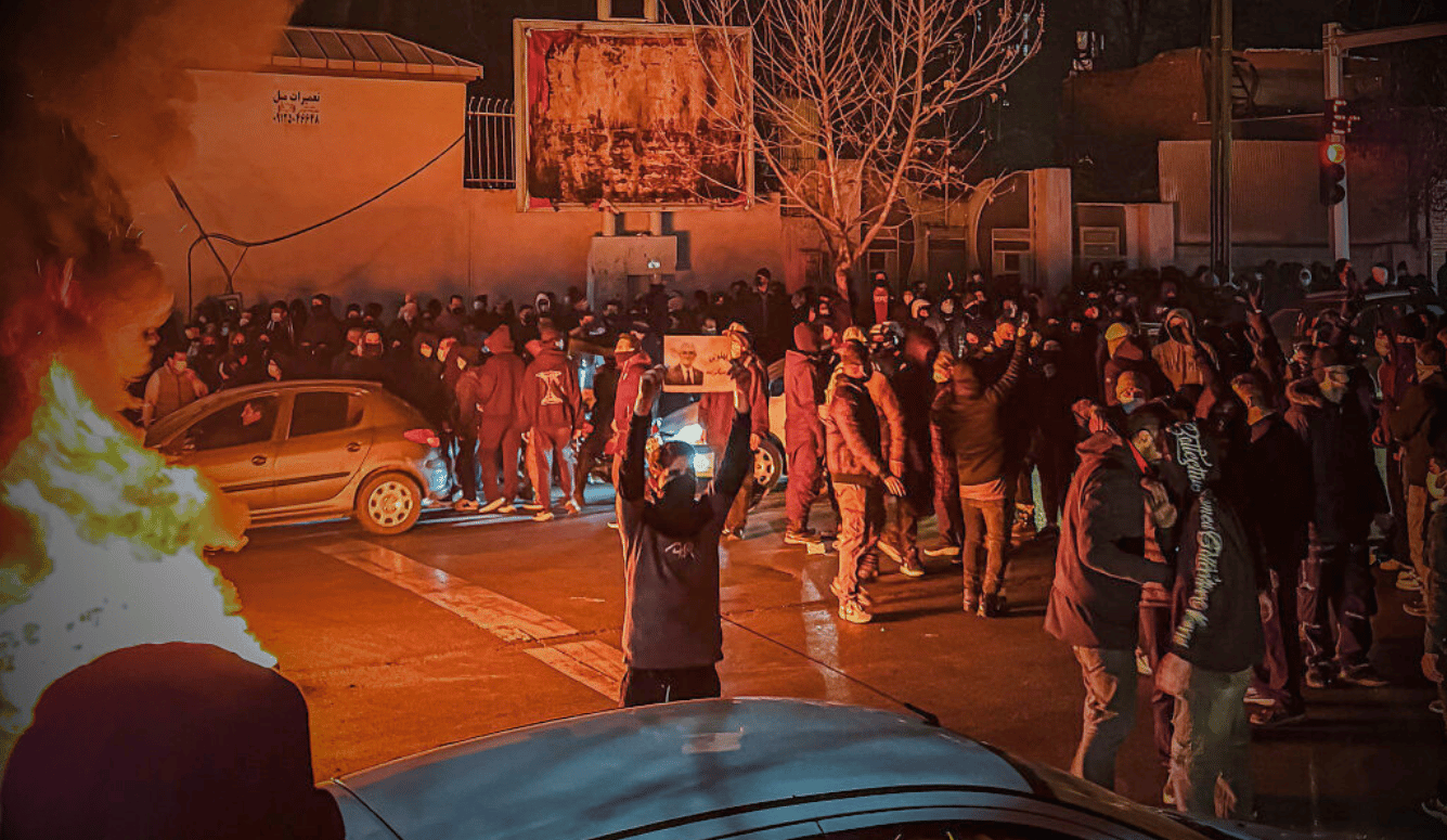 Crowd of masked protesters gathered at night beside burning debris and parked cars on a city street, some holding placards.