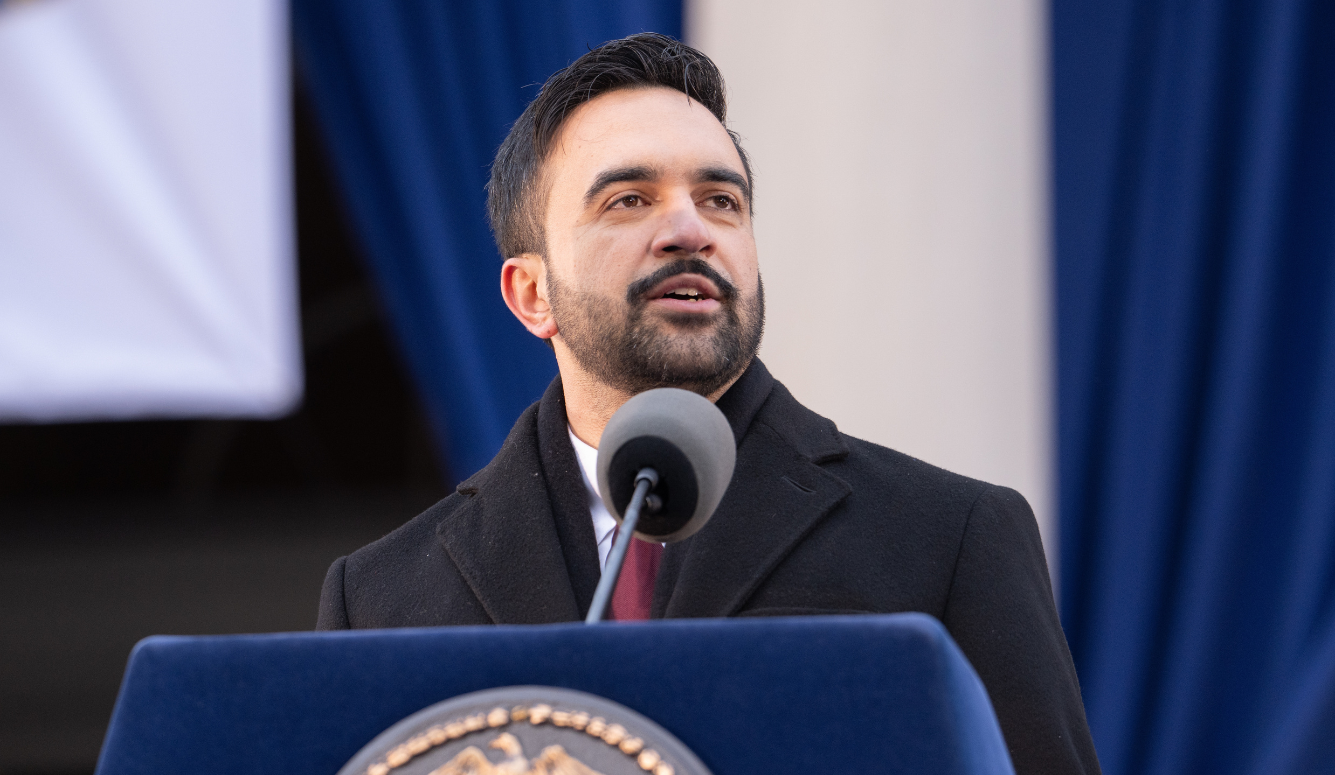 Mamdani is an adult man with short dark hair and trimmed beard speaking at an outdoor podium, wearing a dark coat and tie, with flags behind him.