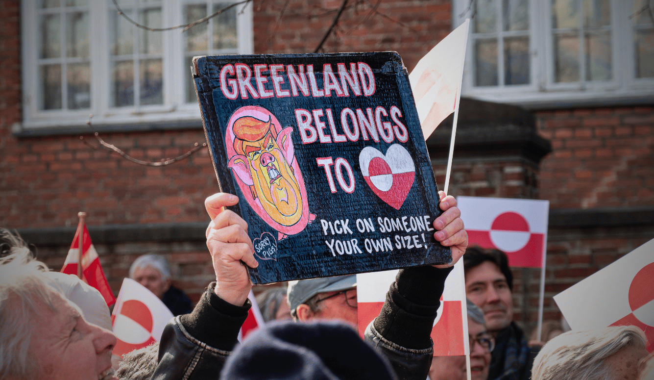 Protester holding placard reading “Greenland belongs to” with cartoon face and heart symbol, during a street demonstration with flags.