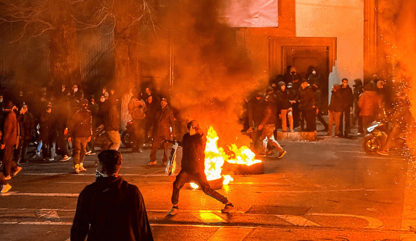 Crowd gathered on a city street at night as a person throws an object towards a burning tyre during a street protest.