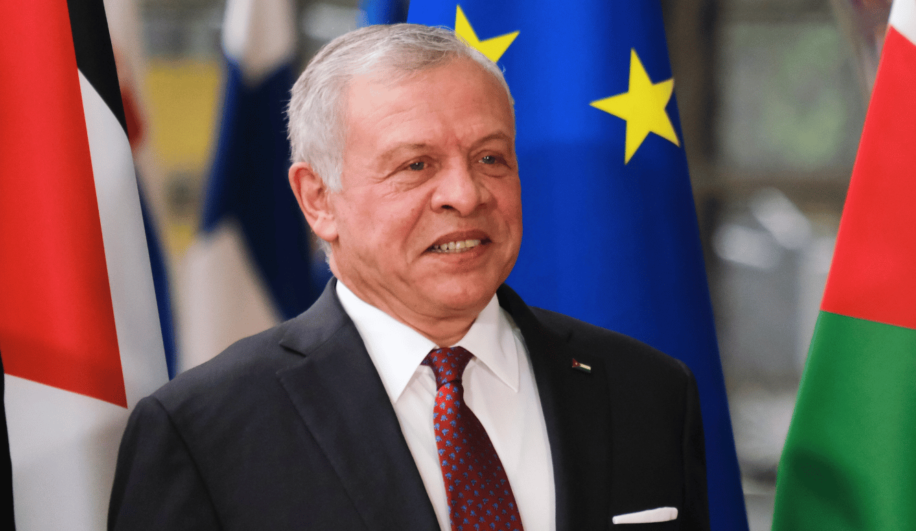 Man in a dark suit and red tie standing before EU and Jordanian flags at a formal diplomatic meeting.