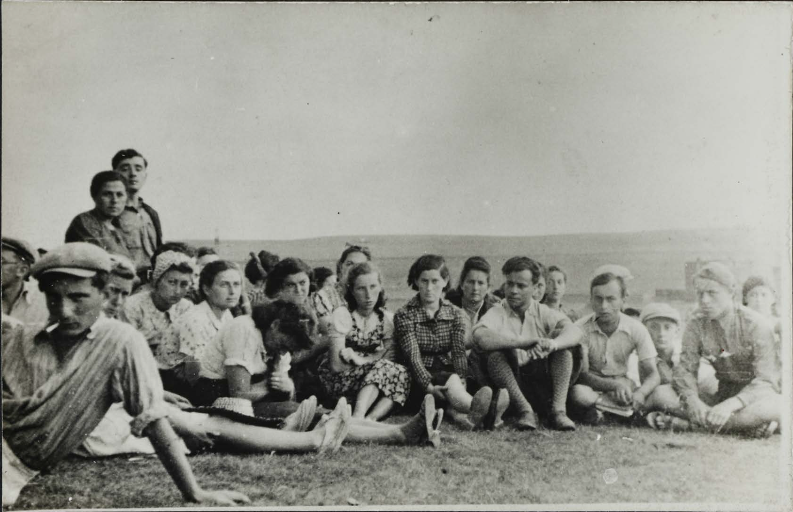 Group of He-Chaluts youth movement members seated on grass outdoors in Będzin, wearing mid-20th-century casual clothing.