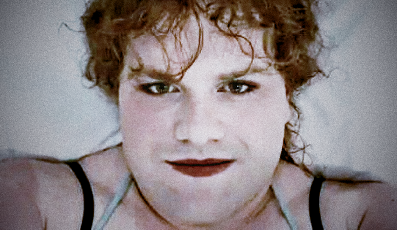 A close-up portrait of a young person with curly reddish-brown hair and fair skin, smiling at the camera against a light-coloured background.