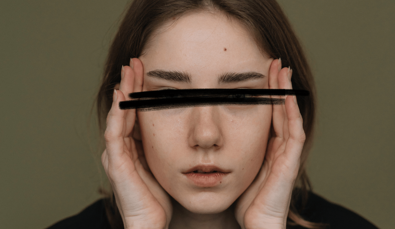 Close-up portrait of young woman holding her temples, eyes obscured by black scribble, against a plain background.