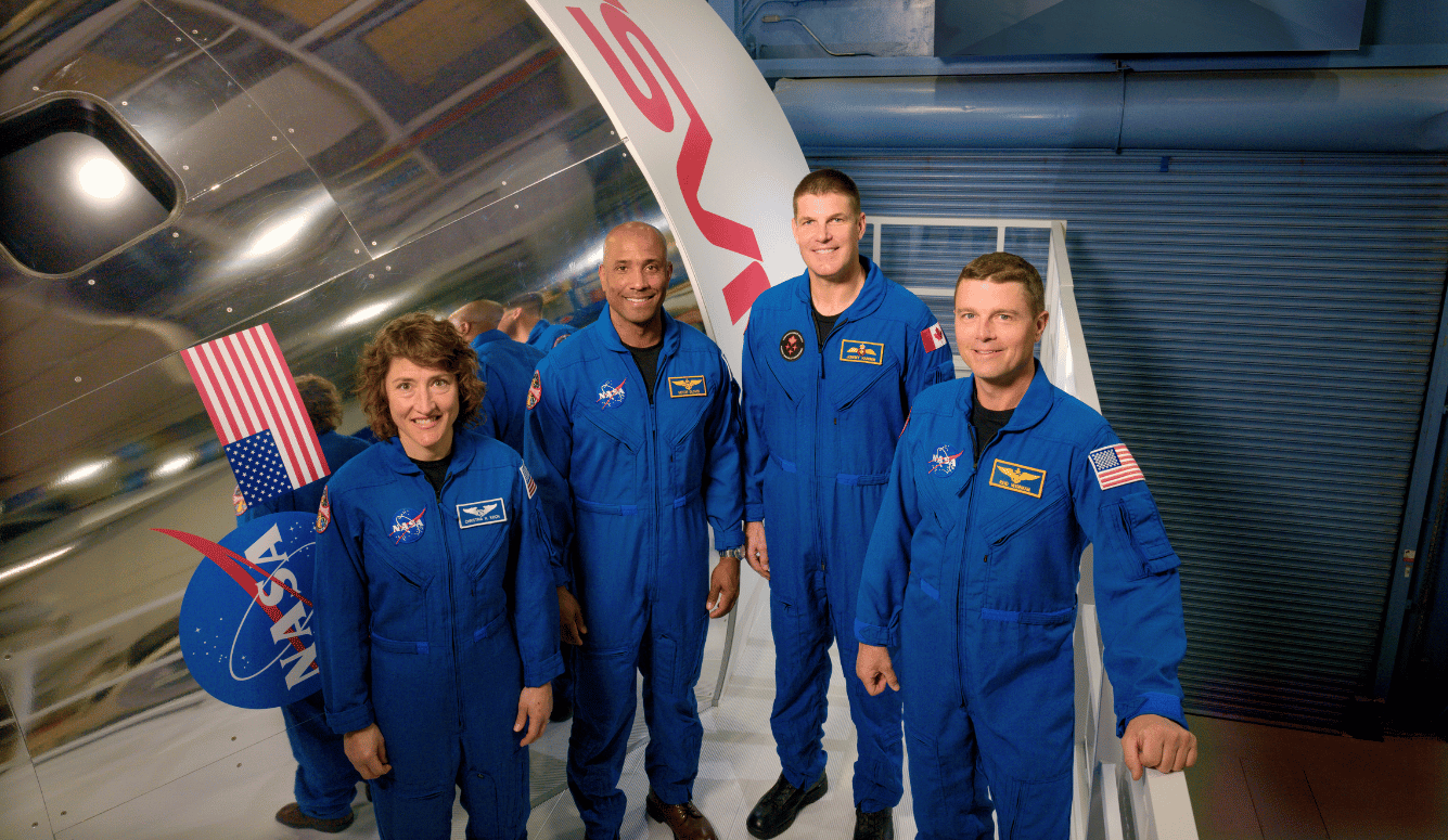 Four NASA astronauts in blue flight suits stand beside a spacecraft mock-up with US flags and NASA logo.