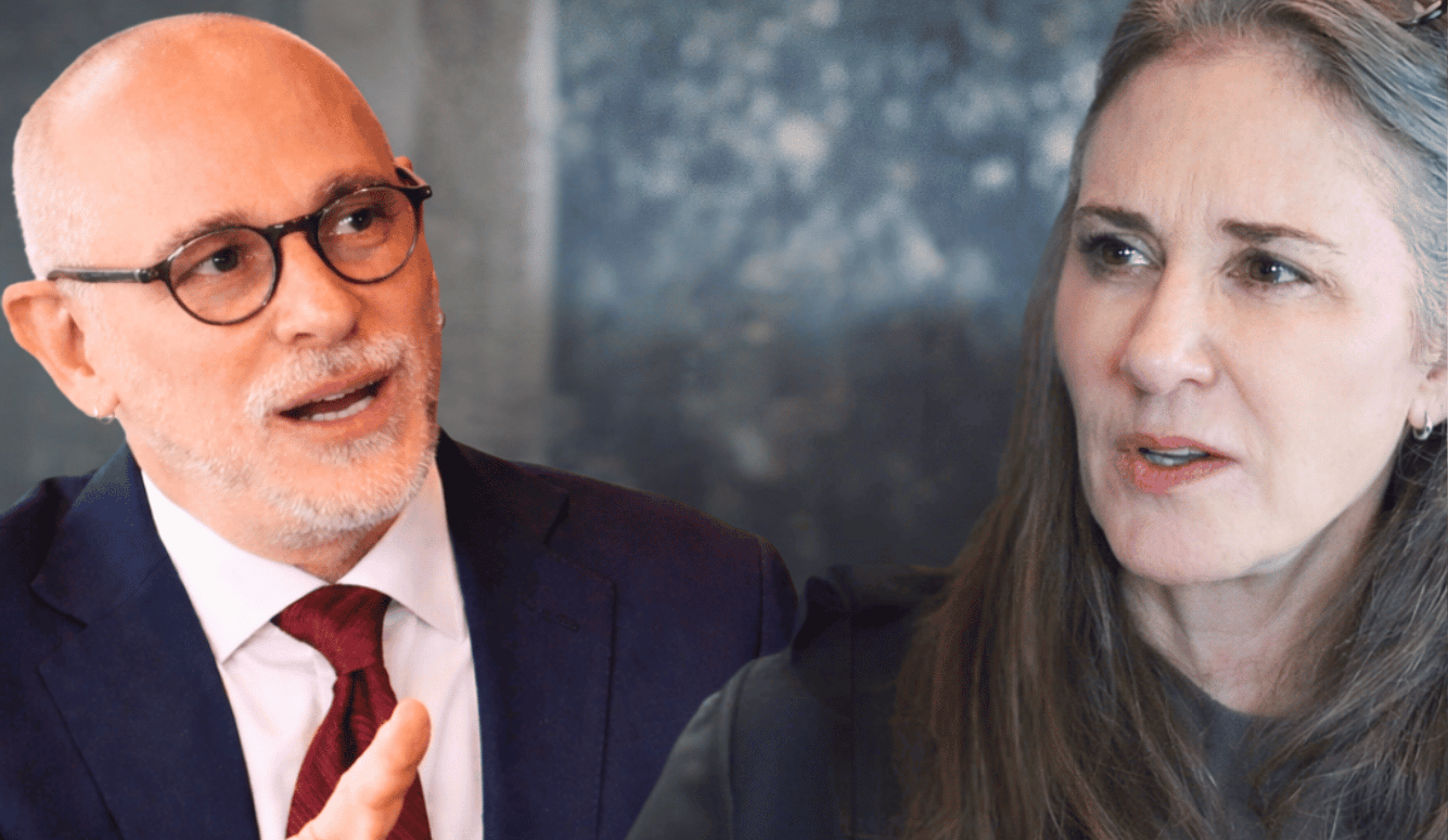 Close-up of a bald man in glasses and suit gesturing as he speaks beside a grey-haired woman indoors.